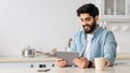 Cheerful arab man using digital tablet while drinking coffee in kitchen at home and checking social networks, copy space Royalty Free Stock Photo