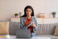 Cheerful Arab woman in headphones using laptop, working or studying online, writing down info during video conference Royalty Free Stock Photo