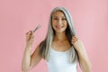 Cheerful Asian lady holds lock of silver hair and scissors on pink background