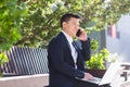 Cheerful asian man businessman working on laptop and talking on the phone, smiling sitting on a park bench near the office on Royalty Free Stock Photo