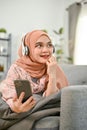 Cheerful Asian Muslim woman relaxing in her living room, listening to music on sofa Royalty Free Stock Photo