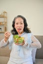 Cheerful Asian 60s aged woman having breakfast in her living room, eating healthy salad Royalty Free Stock Photo