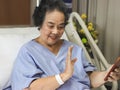 cheerful Asian senior female patient sitting in hospital bed, smiling and looking at mobile phone, video calling and waving Royalty Free Stock Photo