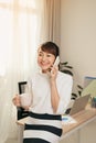 Cheerful Asian woman having conversation on mobile phone and drining coffee at office