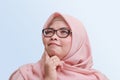 cheerful Asian woman with hijab, standing against blue background, thinking about question with hand on chin Royalty Free Stock Photo