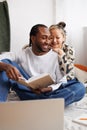 Cheerful asian woman hugging african american boyfriend with book near laptop on bed Royalty Free Stock Photo