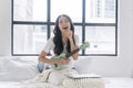 Cheerful asian woman playing ukulele sitting on white bed with pillows beside window at apartment. Joyful young girl holding Royalty Free Stock Photo