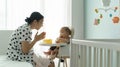 Cheerful Asian Young Mother having fun enjoying while her Little cute baby boy sitting eating on high chair at home. Mom feeding Royalty Free Stock Photo