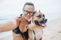 Cheerful asian young woman in eyeglases sitting and doing sekfie with her dog on the beach Royalty Free Stock Photo