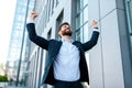 Cheerful attractive young caucasian businessman with beard in suit raises his hands up and rejoices to victory