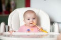 Cheerful baby child eats food itself with spoon. Portrait of happy kid girl in high chair. Royalty Free Stock Photo