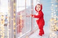 Cheerful baby in red santa`s reindeer costume plays against the background of window and lights of garlands