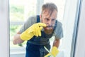 Cheerful bearded man in funny eyeglasses cleans the window with detergents and remove dirt with his finger in gloves.