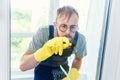 Cheerful bearded man in funny eyeglasses cleans the window with detergents and remove dirt with his finger in gloves.