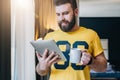 Cheerful bearded man stands and using tablet computer. Guy laughs looking screen of digital tablet while drinking coffee Royalty Free Stock Photo