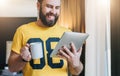 Cheerful bearded man stands and using tablet computer. Guy laughs looking screen of digital tablet while drinking coffee Royalty Free Stock Photo