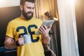 Cheerful bearded man stands and using tablet computer. Guy laughs looking screen of digital tablet while drinking coffee Royalty Free Stock Photo