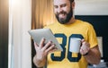 Cheerful bearded man stands and using tablet computer. Guy laughs looking screen of digital tablet while drinking coffee Royalty Free Stock Photo