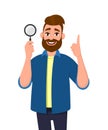 Cheerful bearded young man holding/showing magnifying glass and pointing hand finger up. Search, find, discovery, analyze, inspect