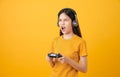 Asian woman in casual yellow t-shirt and playing video games using joysticks with headphones on orange background.