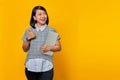 Cheerful beautiful Asian woman holding laptop looking sideways and showing thumbs up sign over yellow background