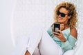 Cheerful beautiful curly young adult woman use mobile phone outdoor sit down on a white garden elegant chair -smile and enjoy the Royalty Free Stock Photo