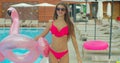 Cheerful beautiful young woman walking by the pool holding sprinkled pink inflatable flamingo float in summer. Pool