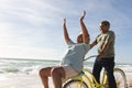 Cheerful biracial senior woman cheering while man riding bicycle at beach on sunny day