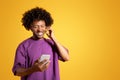 Cheerful black adult curly man in purple t-shirt and wireless headphones looks at phone