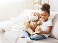 Cheerful black baby girl reading book on couch at home Royalty Free Stock Photo