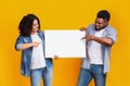 Cheerful black couple holding and pointing at blank advertising board, yellow background Royalty Free Stock Photo