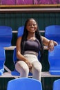 Cheerful black female with long braids sits on blue seat on stadium watching football team training Royalty Free Stock Photo