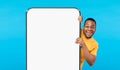 Cheerful Black Guy Peeking Out Behind Big Smartphone With White Blank Screen