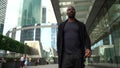 Cheerful black man is walking on street at downtown of modern city, smiling and waving by hand