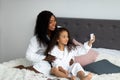 Cheerful black mother with daughter taking selfie in bath robes, posing for photo on bed at home Royalty Free Stock Photo