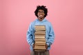 Cheerful black teenager standing with big stack of books, having lots of homework on pink studio background Royalty Free Stock Photo