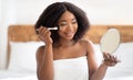 Cheerful black woman applying powder or blusher with brush, using decorative makeup in front of mirror at home Royalty Free Stock Photo