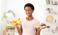 Cheerful black woman holding plates with fruits and desserts indoors, choosing between healthy and junk foods Royalty Free Stock Photo
