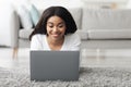 Cheerful black woman relaxing with laptop computer at home, surfing internet while lying on floor in living room Royalty Free Stock Photo