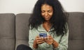 Cheerful black woman sitting on a couch at home and typing a sms message on a smart phone. POC female texting online with a new Royalty Free Stock Photo