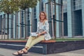 Charming blonde female in modern clothes, studying with a book, sitting on a bench in the park against a skyscraper. Royalty Free Stock Photo