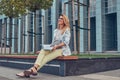 Charming blonde female in modern clothes, studying with a book, sitting on a bench in the park against a skyscraper. Royalty Free Stock Photo