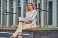 Charming blonde female in modern clothes, studying with a book, sitting on a bench in the park against a skyscraper. Royalty Free Stock Photo