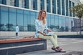 Charming blonde female in modern clothes, studying with a book, sitting on a bench in the park against a skyscraper. Royalty Free Stock Photo