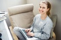 Cheerful blonde in gray home suit with laptop in her hands sitting in chair.