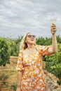 Cheerful blonde woman in bright orange dress with floral print holds glass of champagne high above her head in vineyard Royalty Free Stock Photo