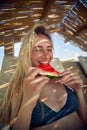 Cheerful blonde woman in swimsuit eating watermelon fruit, feeling blissful. Young woman on summer vacation. Holiday, fun, health