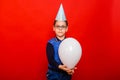 A cheerful boy in a festive cap with a balloon in his hands stands on a red background Royalty Free Stock Photo