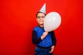 A cheerful boy in a festive cap with a balloon in his hands stands on a red background Royalty Free Stock Photo