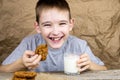 A cheerful boy in a gray T-shirt sits at the table and has breakfast. The child drinks milk and eats cookies